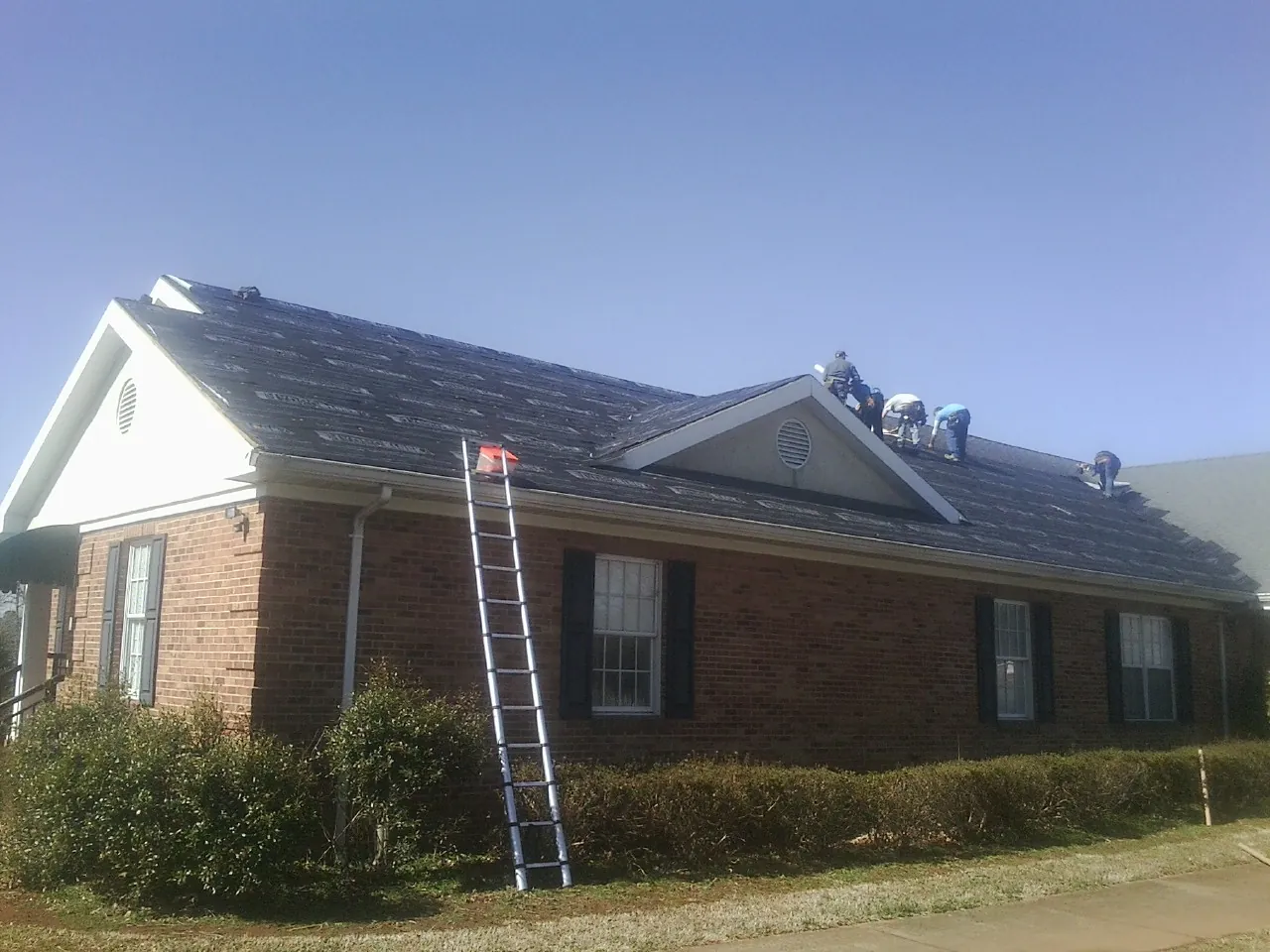 Crew tearing shingles off a brick ranch roof with a ladder set — Emergency Roof Repair work by our crew in Carver