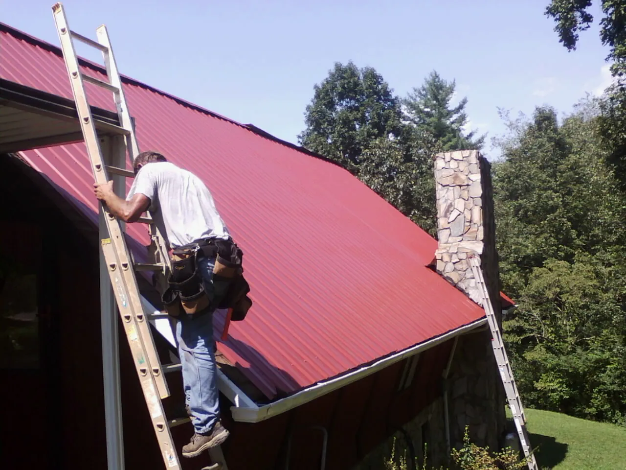 Roofer on a ladder beside a red metal roof and stone chimney — Soffit & Fascia Repair by Roofing Brothers in Carver